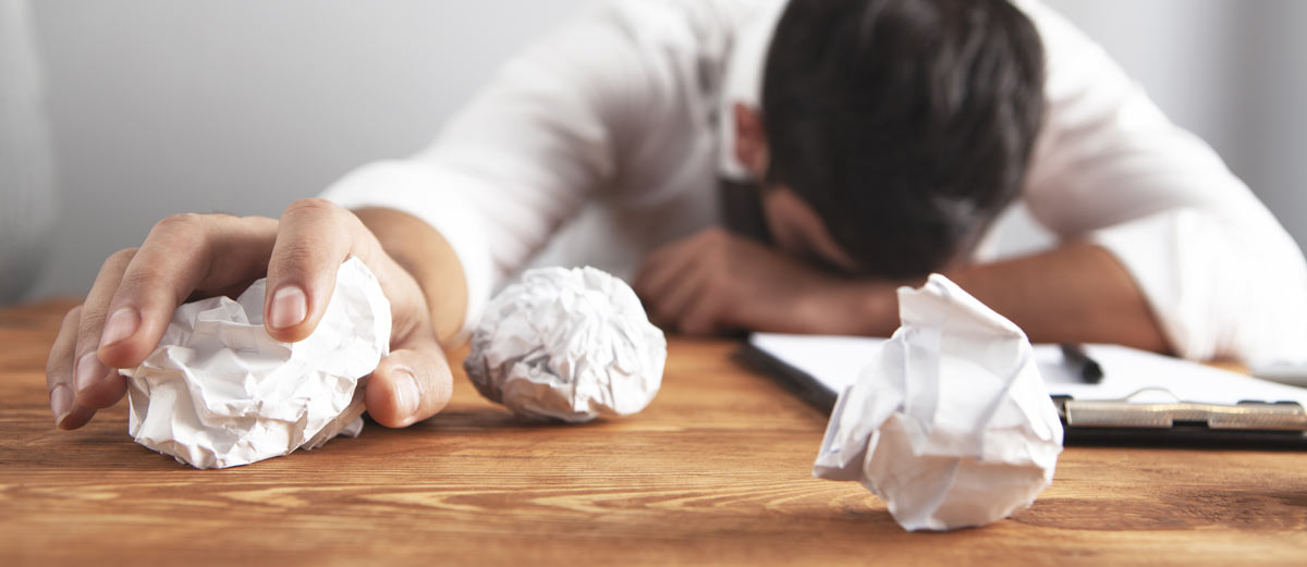 man with head on desk holding crumpled paper in frustration